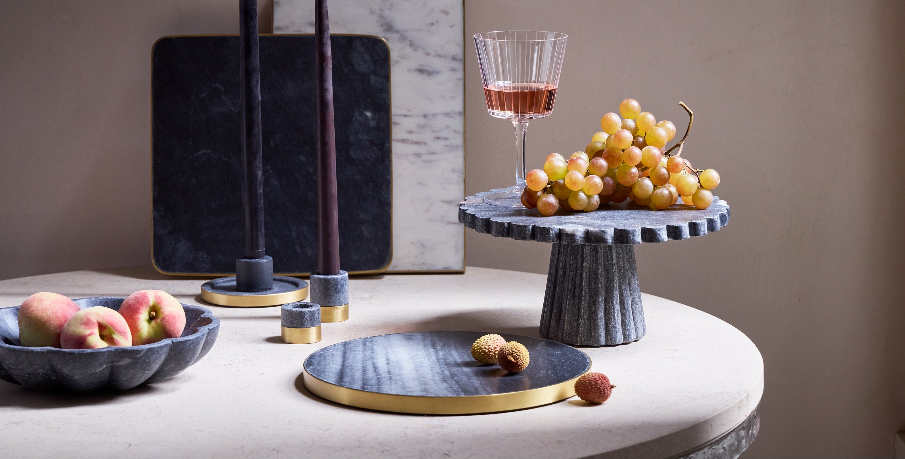 Table setting with black marble plates and saucers, fruit, cake stand, and a glass of red wine on a neutral background. In addition, there are small marble taper candle holders with brass accents.